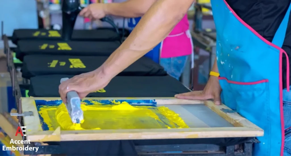 A man in an apron operates a screen printing machine, preparing custom t shirts for printing